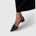 Close-up side view of a black crocodile-embossed patent slingback heel worn with a black midi skirt and silver chain anklet