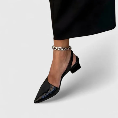Close-up side view of a black crocodile-embossed patent slingback heel worn with a black midi skirt and silver chain anklet