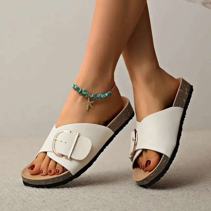 Flat lay of taupe sandals on a woven rug, showing full silhouette with brass buckle and contoured cork footbed