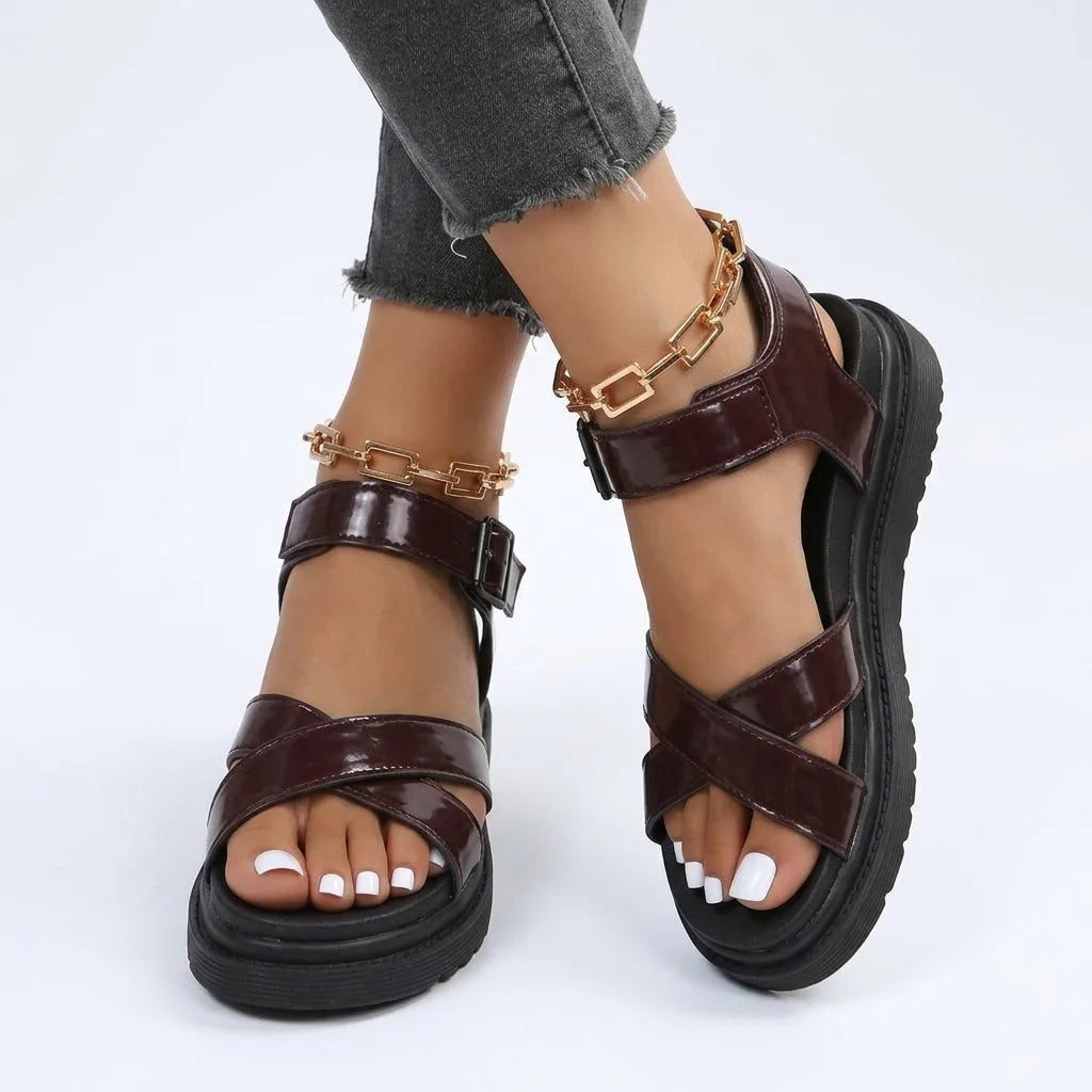 Close-up side view of glossy maroon cross-strap sandals with black platform sole and gold chain anklet