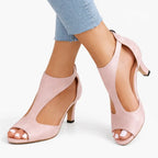Side view of pink suede T-strap heeled sandals with cut-out design, worn on crossed feet with cropped jeans