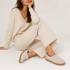 Woman seated wearing beige felt slip-on slippers with burgundy trim while relaxing in a knit outfit