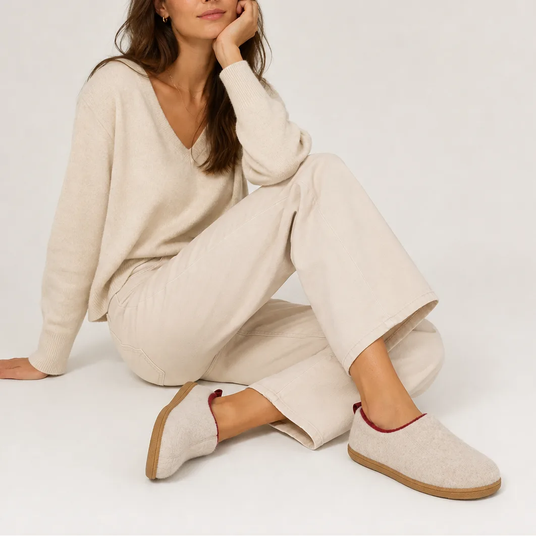 Woman seated wearing beige felt slip-on slippers with burgundy trim while relaxing in a knit outfit