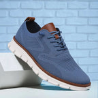 Slate blue knit sneaker against a light blue brick wall with tan leather heel tab and white sole