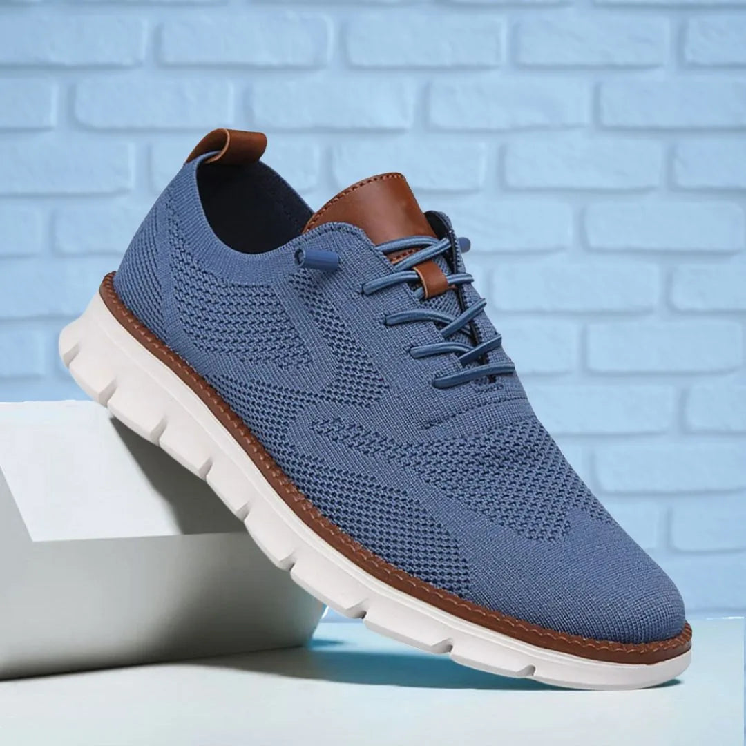 Slate blue knit sneaker against a light blue brick wall with tan leather heel tab and white sole