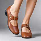 Tan brown leather Mary Jane flats with cut-out side detail and buckle strap, close-up on bare feet