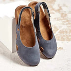 Grey suede slingback flats with round toe, perforated side cut-outs and buckle strap, styled against a textured backdrop.
