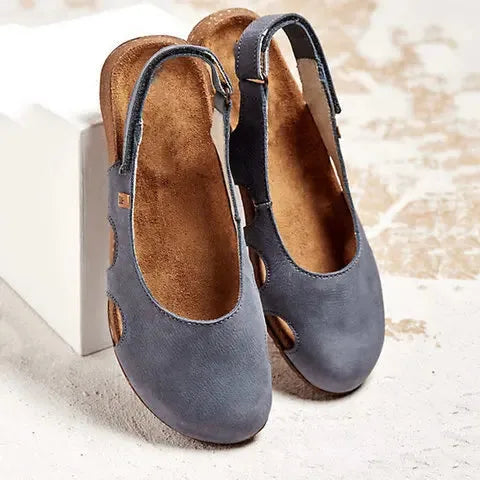 Grey suede slingback flats with round toe, perforated side cut-outs and buckle strap, styled against a textured backdrop.