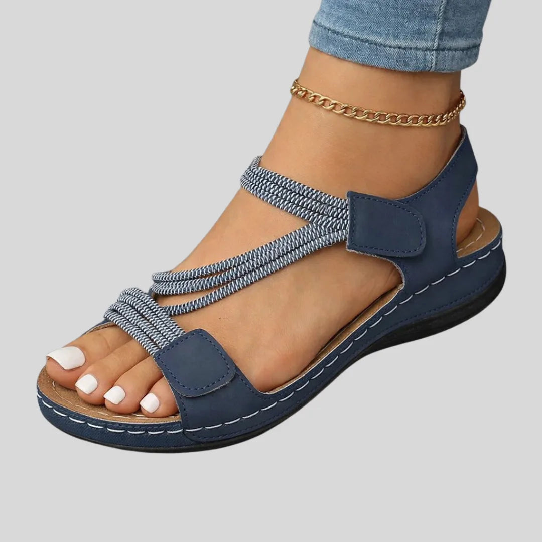 Side view of navy blue strappy sandal with woven elastic crisscross straps and stitched wedge sole, worn with jeans and a gold anklet.