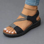 Black strappy wedge sandal with woven textured straps shown on a bent foot, paired with white painted toenails.