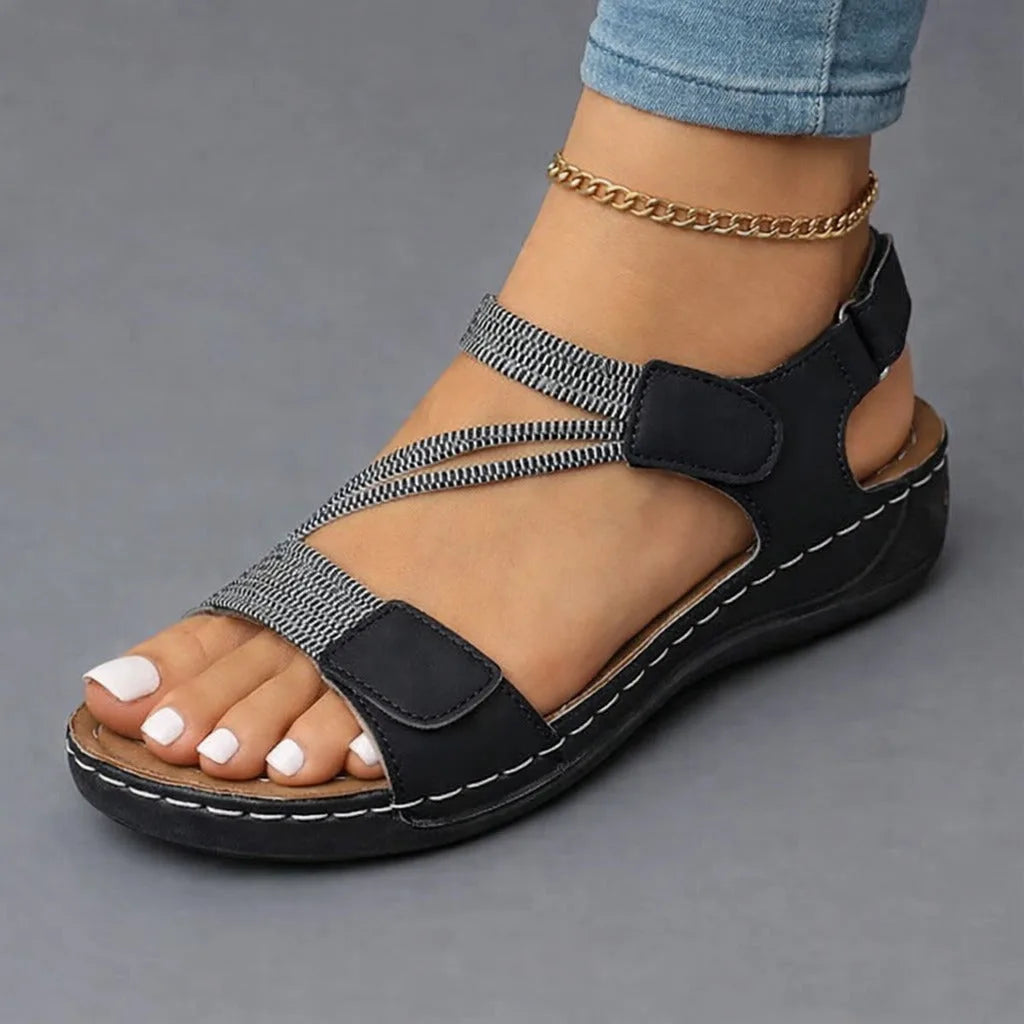 Black strappy wedge sandal with woven textured straps shown on a bent foot, paired with white painted toenails.