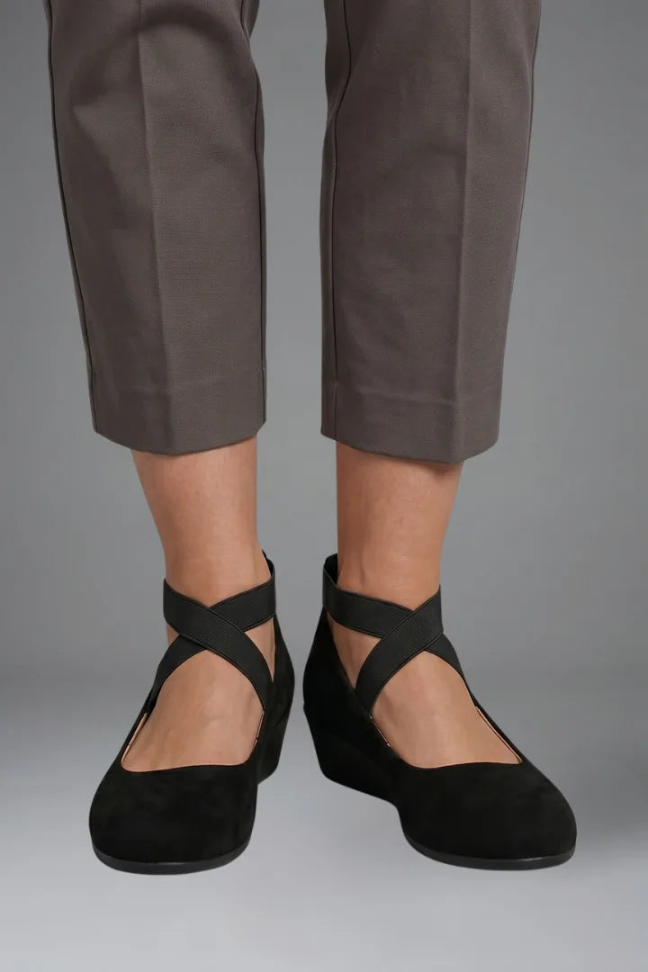 Front view of black suede flats with elastic crossover straps worn with cropped grey trousers