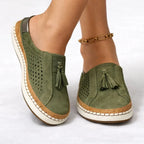 Close-up front view of olive green suede slip-on sneakers with tassel detail worn on bare feet, showing perforated side panels and cork-trim sole