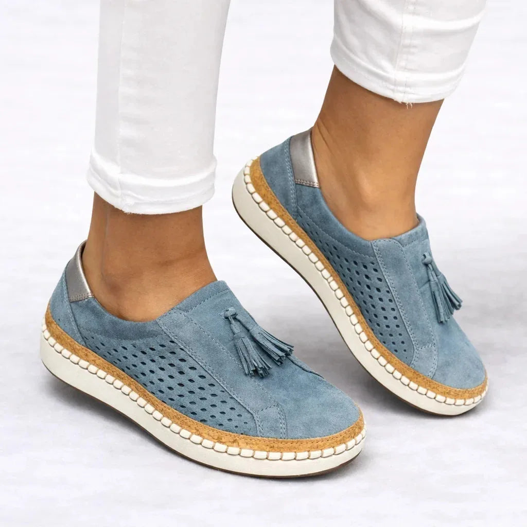 Dusty blue suede slip-on sneakers worn with cuffed white jeans, showing the tassel trim and metallic heel panel