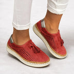 Brick red suede slip-on sneakers worn with white jeans, showing the tasselled vamp and bronze-tone heel accent