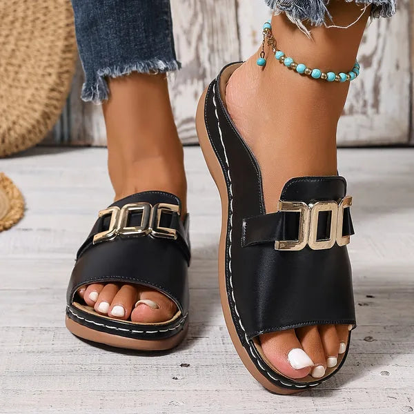 Close-up side view of black faux leather slide sandals with gold buckle detail and white stitched trim, worn with denim and a turquoise beaded anklet