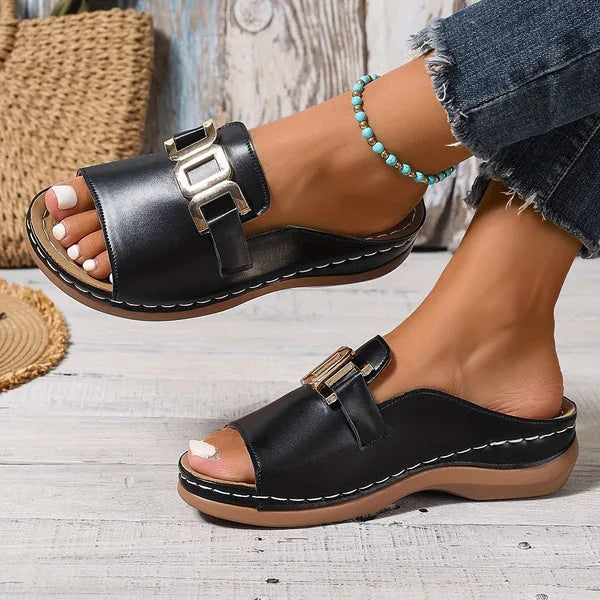 Top-down view of black slide sandals with gold buckle worn outdoors on wood decking, paired with a turquoise beaded anklet