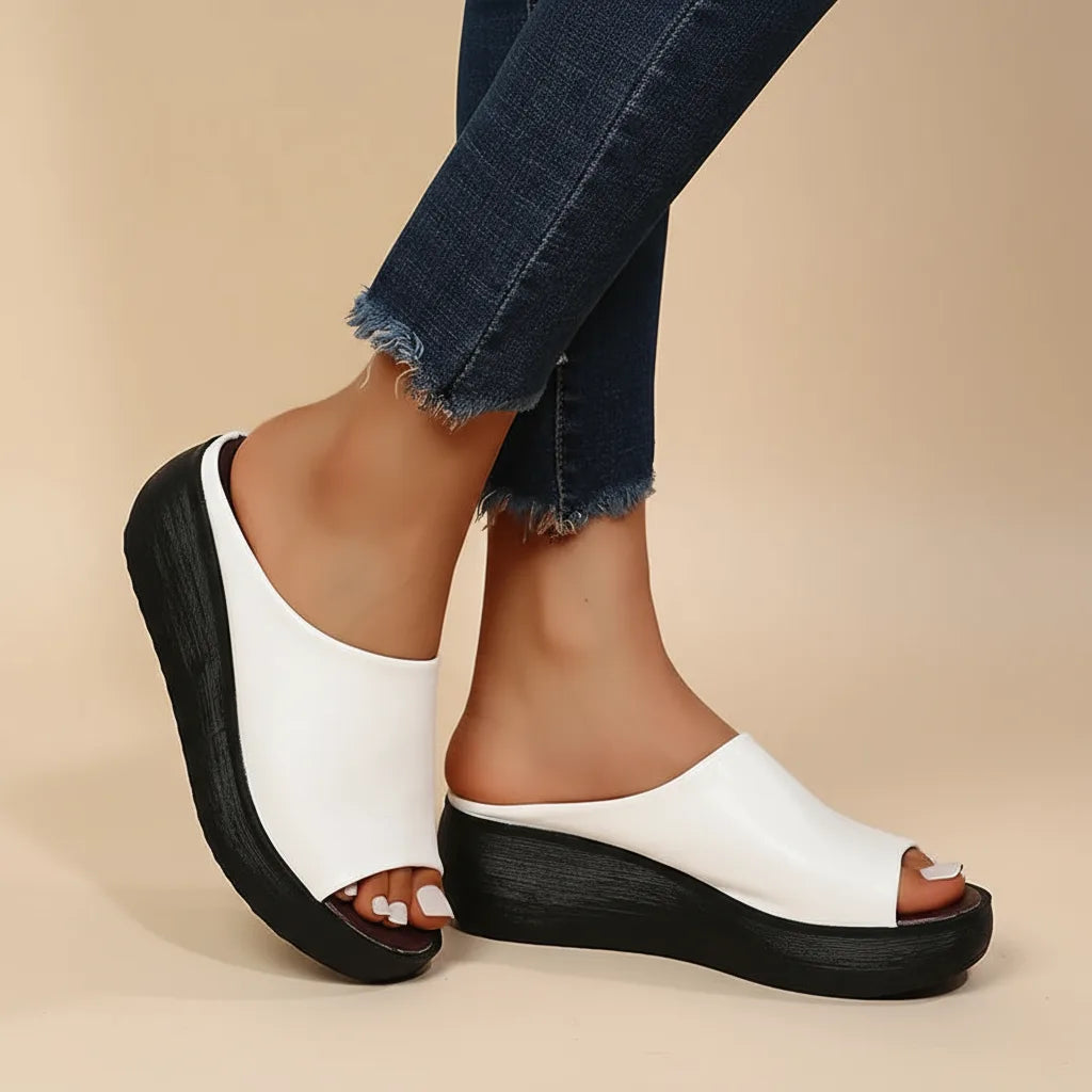 Side view of white faux leather peg-toe wedge slide with thick black platform sole, worn with cropped jeans