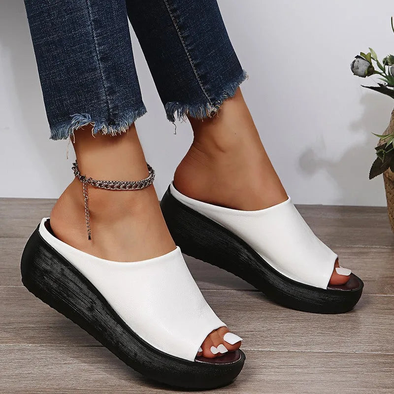 Close-up front view of white peep-toe wedge slides on both feet beside a woven basket