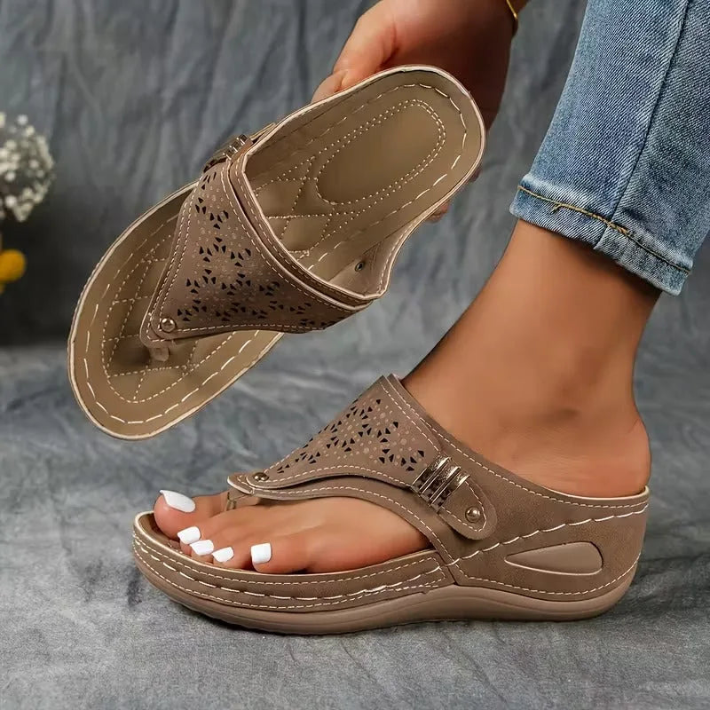 Taupe grey wedge sandal with perforated strap and cushioned footbed, displayed on hand next to denim jeans