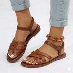 Side view of brown woven strappy flat sandal with elastic ankle strap, worn with rolled-up jeans and a chain anklet