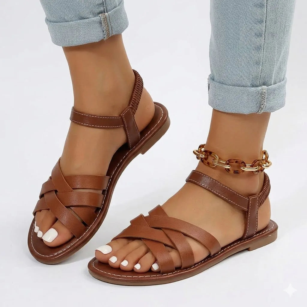 Side view of brown woven strappy flat sandal with elastic ankle strap, worn with rolled-up jeans and a chain anklet
