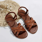 Brown woven flat sandals styled on bare feet next to a palm leaf and pebble decor, showing side profile