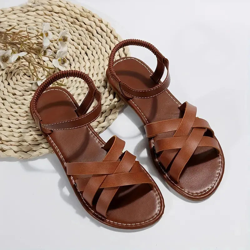 Brown woven flat sandals styled on bare feet next to a palm leaf and pebble decor, showing side profile