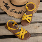 Pair of black crisscross strap flat sandals with elastic ankle strap placed on wooden slats beside a straw hat
