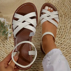 White crisscross strap flat sandals worn on feet, seated on a woven straw mat with white flowers nearby