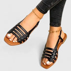 Black woven strappy flat sandals with slingback strap, shown worn alongside cropped dark denim jeans.