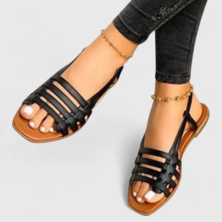 Black woven strappy flat sandals with slingback strap, shown worn alongside cropped dark denim jeans.