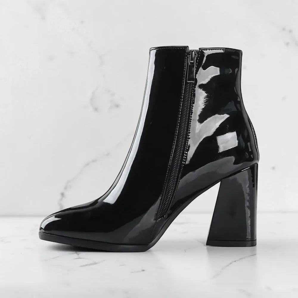 Side profile of a black patent leather ankle boot with side zip and block heel on a marble backdrop.
