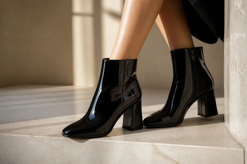 Woman crossing her legs while wearing black patent ankle boots with a chunky block heel in sunlight.