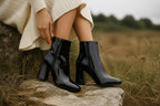 Detail shot of black patent ankle boots with visible zip pull against a natural grassy setting.
