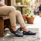Man seated on a wooden bench wearing navy perforated leather loafer-sandals with cobblestone street in background