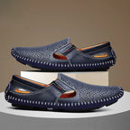 Pair of navy perforated leather slip-on shoes stacked on a white block against a warm brown studio background