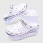 White clogs with iridescent silver heel strap and chunky platform, one shoe resting against a white box