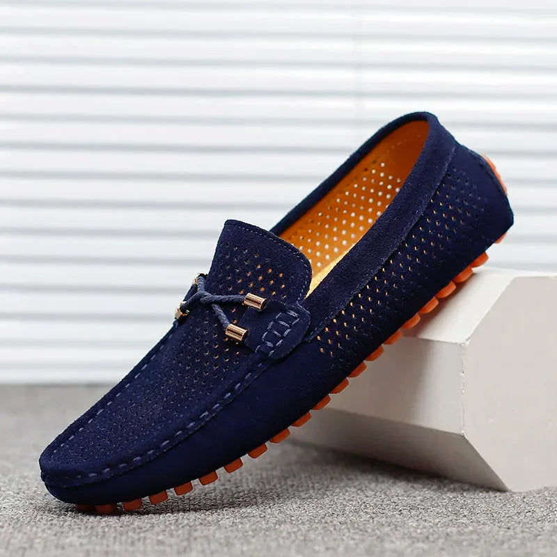 Navy suede perforated loafer with bow lace detail and orange sole, resting against a white block on grey flooring