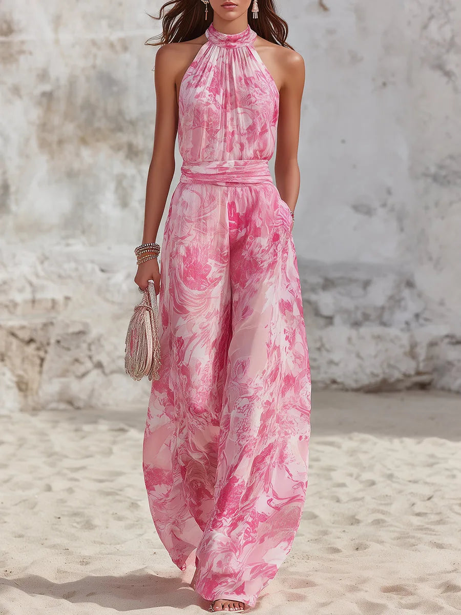 Woman walking a sandy beach in a pink marble-print halter-neck jumpsuit with wide-leg trousers and a matching tie waist