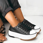 Black lace-up platform sneaker with white toe cap and contrast stitching, worn with black trousers and plaid coat