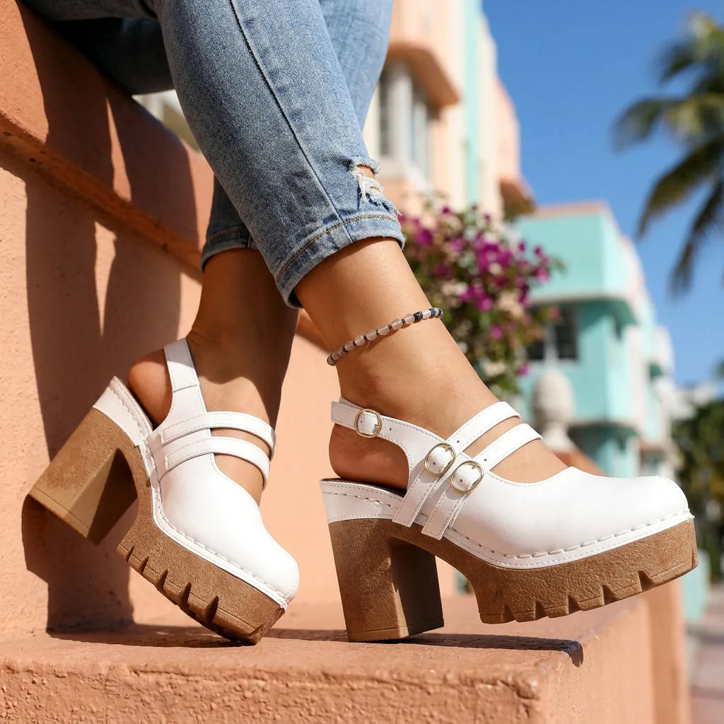White platform clog heels with double strap buckle detail, worn sitting on a pastel-coloured ledge