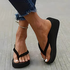 Side view of a black platform flip-flop with thick sole, worn on bare feet with cuffed denim jeans and a chain anklet