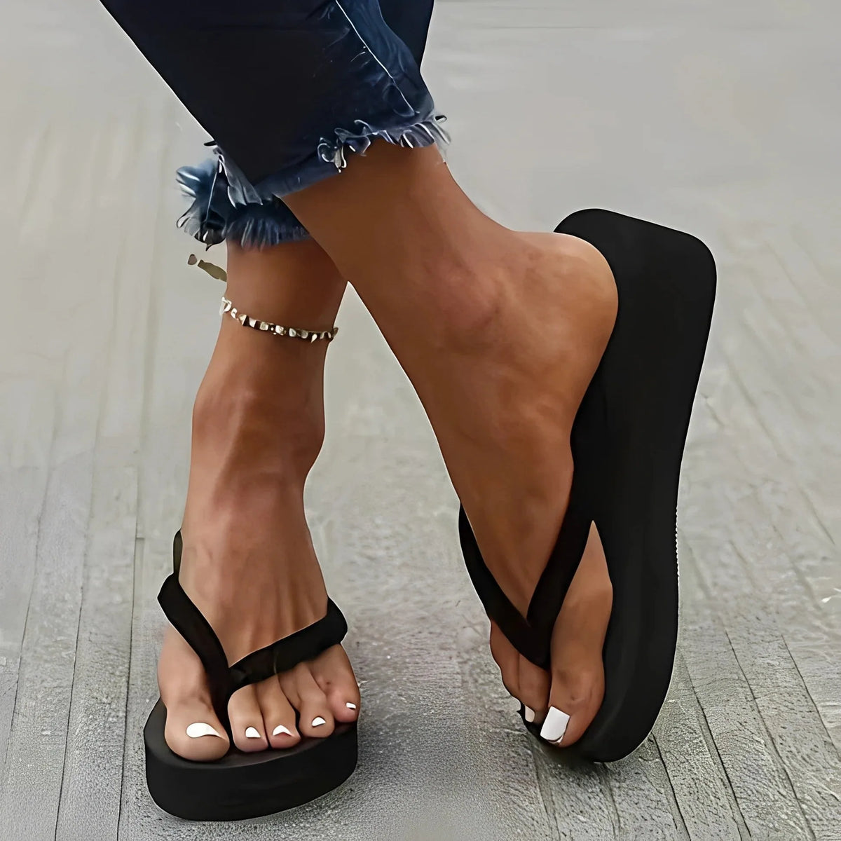 Side view of a black platform flip-flop with thick sole, worn on bare feet with cuffed denim jeans and a chain anklet