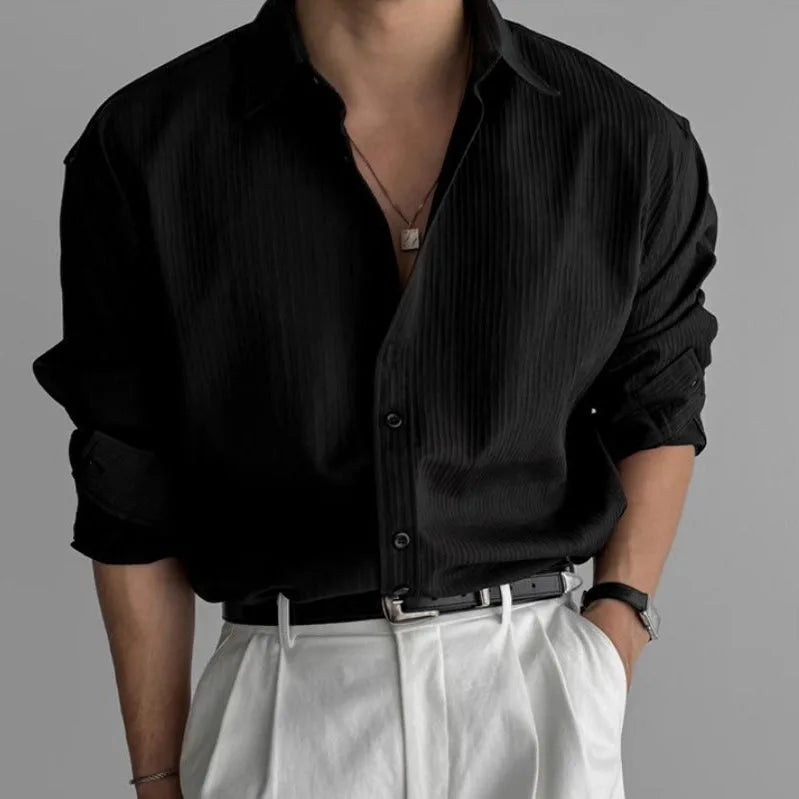 Man wearing an open-collar black pleated textured shirt layered with a silver necklace over white trousers