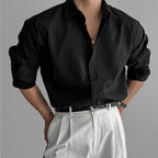 Side angle of a man in a black pleated textured shirt with rolled sleeves and a black leather belt