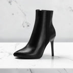 Side profile of a black leather ankle boot with pointed toe and thin stiletto heel on a marble surface