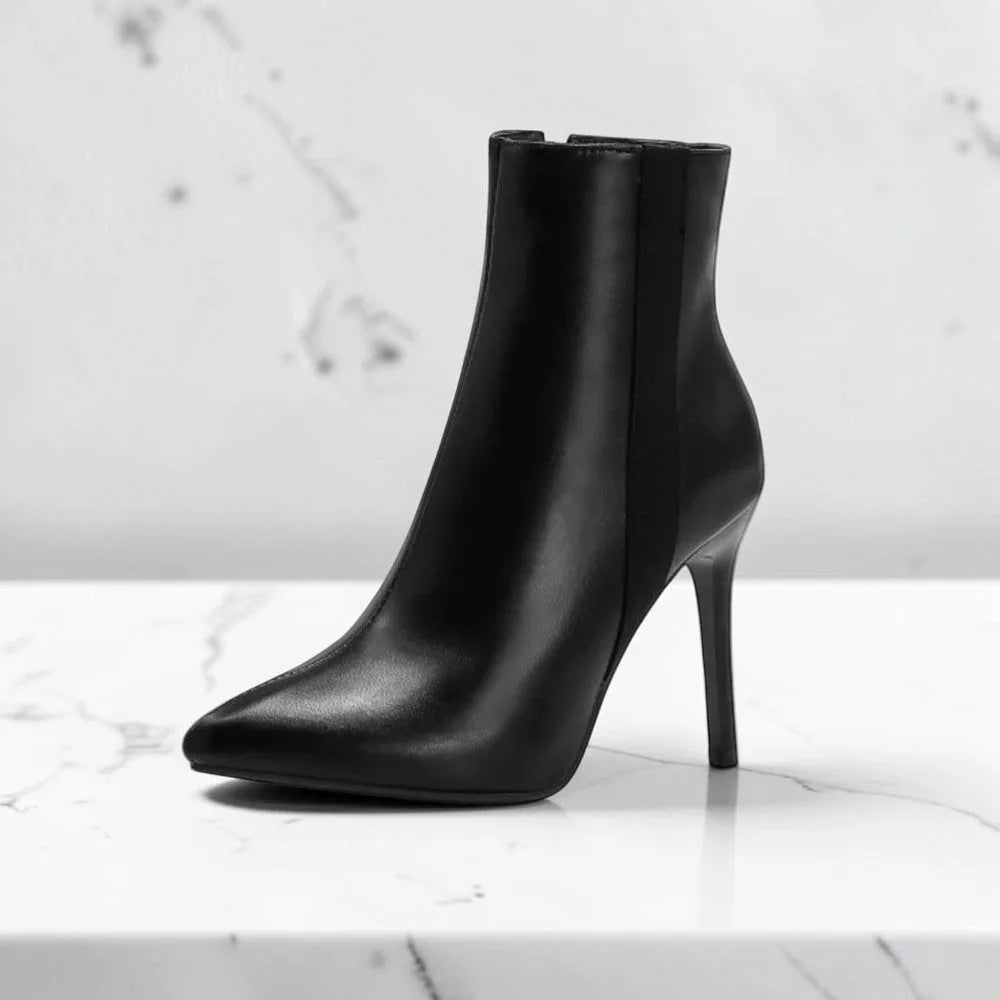 Side profile of a black leather ankle boot with pointed toe and thin stiletto heel on a marble surface