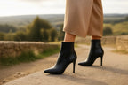 Close-up side view of black leather ankle boots showing the zip closure and slender heel on a stone path