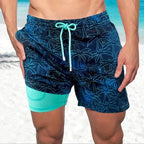 Model standing on a sandy beach wearing navy palm print swim shorts with teal colour-block hem and compression lining